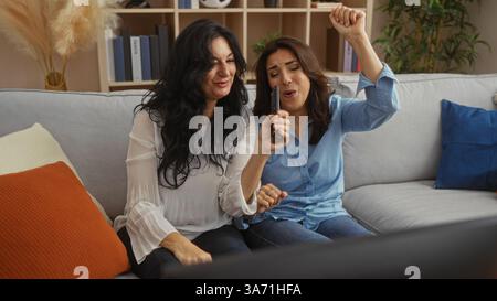 Due donne cantano e ballano gioiosamente in salotto utilizzando il telecomando come microfono, mostrando una scena di amicizia e divertimento a casa. Foto Stock