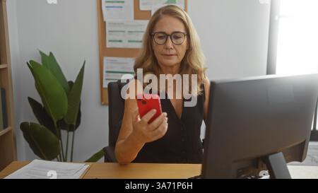 Donna che lavora in ufficio che controlla lo smartphone, donna bionda matura alla scrivania con computer in ambiente professionale, ambiente interno con documenti e p Foto Stock