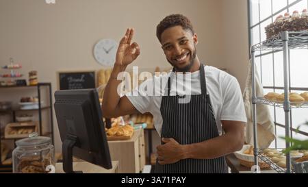 Giovane sorridente con un gesto discreto, in piedi in una panetteria con pane e dolci sullo sfondo, indossando un grembiule nero all'interno in un caldo e luminoso Foto Stock