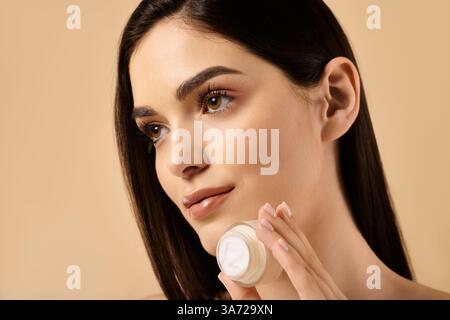 Una giovane donna brillante mette in risalto il suo aspetto impeccabile tenendo delicatamente in mano il prodotto per la cura della pelle. Foto Stock