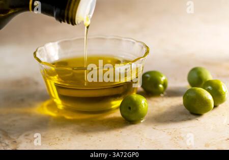 Olio d'oliva versato in un piccolo recipiente di vetro con le olive sul tavolo Foto Stock