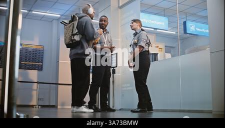 Affollato terminal dell'aeroporto internazionale: Due agenti di sicurezza multietnici in uniforme che parlano, in piedi al checkpoint di sicurezza, mostrando al passeggero maschio la strada per la porta d'imbarco per il suo volo in aereo. Foto Stock
