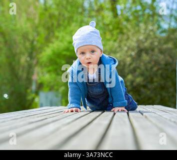 Ragazzo fino a un anno di età, aspetto europeo, occhi blu si siede in estate su una panchina di legno vestita con tute in denim e un berretto leggero. Alta qualità Foto Stock