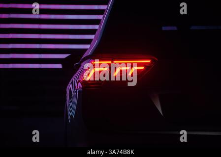 Luce di coda di un'auto moderna di notte. Immagine ravvicinata della luce posteriore a LED illuminata di sinistra, linee al neon viola sul retro. Riflesso delle luci della città. Foto Stock