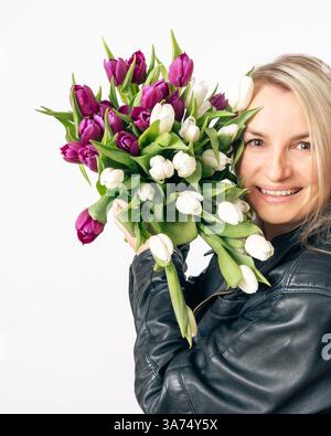 Una donna bionda allegra con una giacca in pelle nera racchiude un grande bouquet di tulipani bianchi e viola. Sorride mentre guarda i fiori contro un Foto Stock
