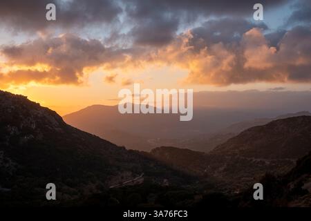 Ecco una didascalia per l'immagine: **L'ora d'oro bagna il paesaggio montuoso con un caldo bagliore mentre il sole tramonta, dipingendo le nuvole con sfumature di oranghi Foto Stock