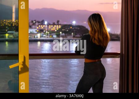 Una giovane donna si erge sul balcone di una cabina di una nave da crociera che guarda il porto e lo skyline di Puerto Vallarta, in Messico di notte. Foto Stock