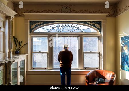 Un maschio adulto anziano sta penosamente davanti a una finestra sporca nel salotto di una casa vittoriana con un gatto che dorme su una sedia accanto. Foto Stock