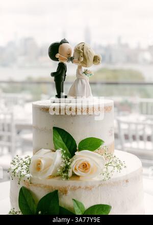 Una torta nuziale splendidamente decorata con due statuette e adornata con fiori freschi, ambientata in un luogo panoramico. Foto Stock