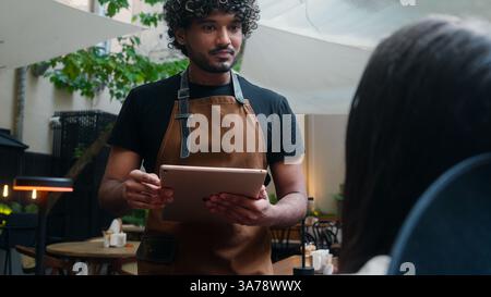 Un uomo indiano sorridente, un ragazzo arabo latino-americano, un cordiale barista del caffè in grembiule, chef, caffè, servizio clienti, ordine del cliente, cibo, pranzo e cena Foto Stock