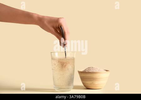 Mano femminile con cucchiaio e buccia di psico in un bicchiere d'acqua su sfondo beige Foto Stock