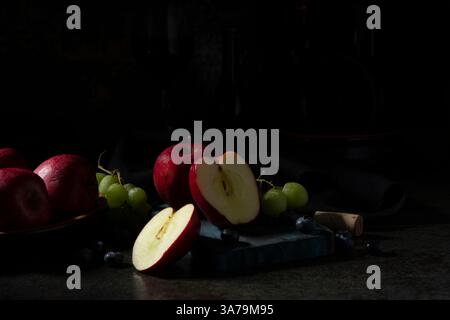Mele rosse, uva verde e mirtilli su un tagliere su un tavolo in ceramica nera con bottiglie di vino sullo sfondo scuro Foto Stock