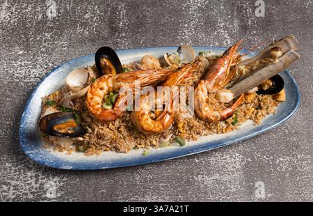Gusta un delizioso piatto di riso fritto a base di pesce con gamberi, cozze, vongole e vongole di rasoio, servito su un elegante piatto blu Foto Stock