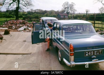 Una donna siede in un Austin Cambridge parcheggiato su un vialetto rurale britannico durante gli anni '1960 La classica berlina blu, un popolare veicolo per famiglie dell'epoca, è circondata da campagna e da una pila di mattoni, forse accennando alla costruzione o al rinnovamento in corso. Foto Stock