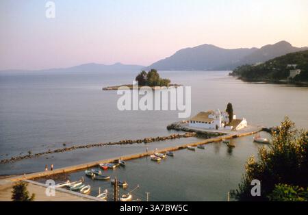 Una vista serena del Monastero di Vlacherna, situato su un piccolo isolotto al largo della costa di Corfù, Grecia. Catturato durante la luce soffusa del 1970, il monastero si trova pacificamente alla fine di una stretta strada rialzata, con piccole barche ormeggiate nelle vicinanze e Pontikonisi (Isola del mouse) visibile sullo sfondo. Un'immagine senza tempo di solitudine spirituale e bellezza ionica. Foto Stock