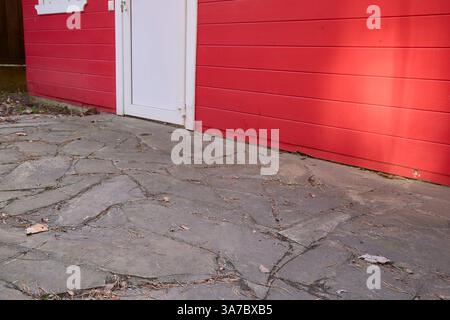 Il patio in pietra conduce ad una vibrante parete di legno rosso con una porta bianca che mostra un ambiente rustico all'aperto. L'area è cosparsa di foglie secche, creando un Foto Stock
