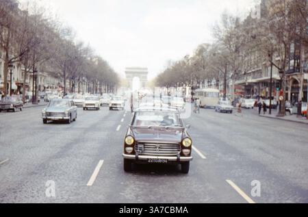 Una classica scena degli anni '1960 lungo gli Champs-Élysées di Parigi, in Francia, caratterizzata da un traffico intenso diretto verso l'Arco di Trionfo. Un taxi Peugeot di colore scuro conduce il flusso di veicoli d'epoca lungo l'ampio viale alberato. Foto Stock