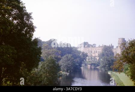 Castello di Warwick visto dall'altra parte del fiume Avon, circondato da alberi maturi e riflesso nell'acqua. Una tranquilla scena che cattura la grandezza della fortezza medievale in una giornata nebulosa negli anni '1960 Foto Stock