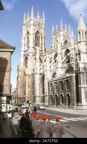 Una vista della Cattedrale di York nel giugno 1965, che mostra le sue grandi torri gotiche e le decorazioni in pietra. A York, Inghilterra, la cattedrale è formalmente conosciuta come la Cattedrale e la Chiesa Metropolita di San Pietro. In primo piano, un gruppo di scolari con uniformi e berretti rossi abbinati passeranno davanti all'iconica struttura Foto Stock