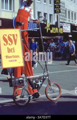 Un vivace artista di strada su palafitte pubblicizza una vendita stagionale presso i grandi magazzini Horten di Hildesheim, Germania, durante l'estate del 1986. Vestito con tute di rosso brillante, l'intrattenitore cavalca una bicicletta alta comicamente modificata lungo una trafficata strada pedonale per lo shopping, catturando l'attenzione con un cartello giocoso e un'esibizione stravagante Foto Stock