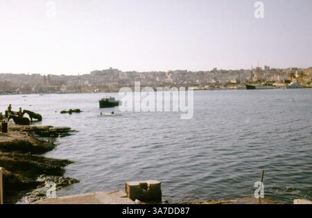 Una vista degli anni '1960 attraverso il Grand Harbour di Malta, con nuotatori e piccole barche in primo piano e lo storico skyline di Senglea e Vittoriosa (parte delle tre città) che si innalzano in lontananza, catturato su una pellicola da 35 mm. Foto Stock