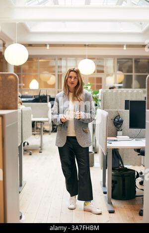 Ritratto a tutta lunghezza di una donna sicura di sé che tiene la tazza mentre si trova in ufficio Foto Stock
