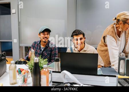 Amici maschi sorridenti seduti in classe mentre studiano all'università Foto Stock