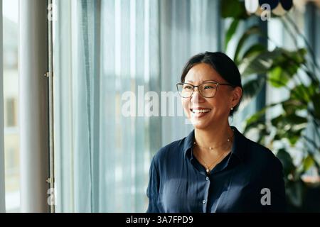 Donna d'affari felice e matura con occhiali che guarda dalla finestra dell'ufficio Foto Stock