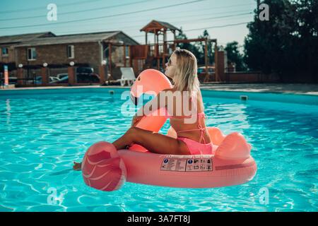 Una donna è seduta su un galleggiante rosa di fenicottero in una piscina. La scena è luminosa e allegra, con la donna che si gode il suo tempo in acqua. Foto Stock