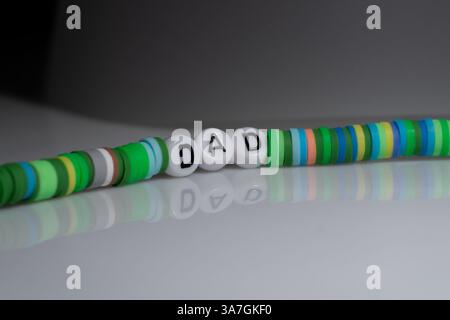 Immagine ravvicinata del braccialetto con perline colorate che scrive IL PAPÀ su sfondo riflettente con superficie lucida. Foto Stock