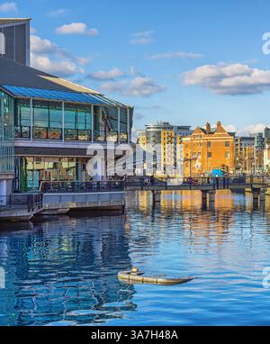 Un moderno centro commerciale costruito in vetro si trova accanto ad un vecchio molo con un ponte sull'acqua. Altri edifici sono sullo sfondo e un cielo W. Foto Stock