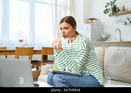 Una bella giovane donna si siede attentamente a casa, concentrandosi sul suo studio online mentre prende appunti. Foto Stock