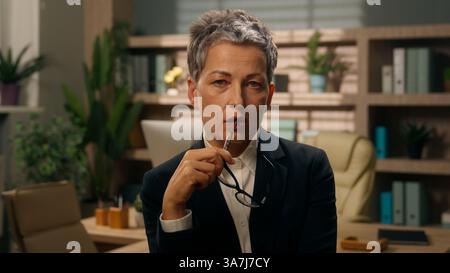Grave penosa donna d'affari caucasica vecchia donna d'affari anziana donna matura datore di lavoro in pensione in ufficio che guarda la fotocamera che tiene gli occhiali dentro Foto Stock