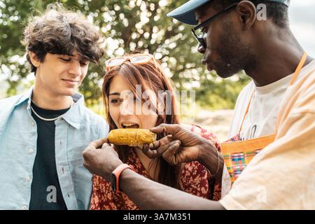 Gruppo multiculturale di giovani amici che condividono il mais alla griglia durante il barbecue all'aperto in una giornata estiva di sole, godendosi un momento di allegria e di allegria insieme Foto Stock