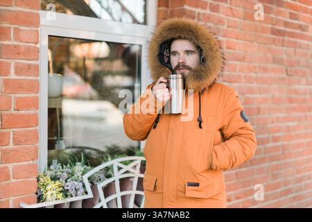 Uomo con cappotto invernale che beve da thermos sulla strada della città invernale vicino alla vetrina del negozio. Comfort, routine e calore durante una fredda giornata urbana all'aperto. Copia Foto Stock