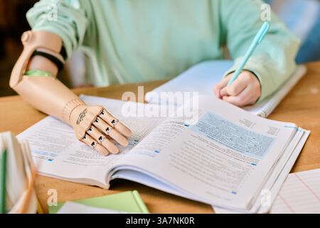 Persona con braccio protesico che studia e prende appunti a una scrivania piena di libri aperti e cancelleria. Sottolineando l'uso di dispositivi assistivi in ambito educativo Foto Stock