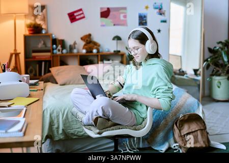 Individuale con braccio protesico che studia in una camera accogliente con cuffie e computer portatile, circondato da scrivania, libri, ornamenti e illuminazione soffusa Foto Stock