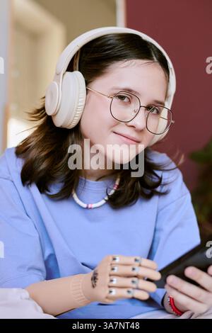 Ritratto di una bellissima ragazza adolescente con una protesi che tiene lo smartphone e sorride in un ambiente rilassato con uno sfondo accogliente Foto Stock