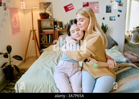 La madre amorevole abbraccia delicatamente la figlia con il braccio protesico mentre si siede sul letto in una camera accogliente e personalizzata circondata da piante, decorazioni e libreria che espone vari oggetti Foto Stock