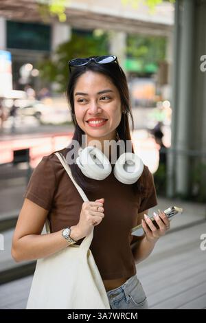 Giovane turista asiatico che cammina attraverso una vivace strada di bangkok, tenendo in mano uno smartphone, indossando le cuffie e portando con sé una borsa tote alla moda, sorridente Foto Stock