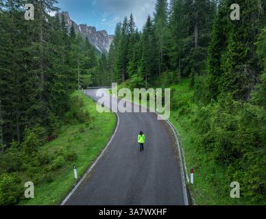 Donna che cammina su una tortuosa strada di montagna nella lussureggiante foresta verde in estate giorno piovoso. Dolomiti, Italia. Veduta aerea della ragazza, strada alpina in pas di montagna Foto Stock