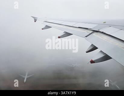 Ali di aeroplani che volano sopra un aeroporto nebbioso durante un atterraggio, con altri aerei parcheggiati a terra Foto Stock