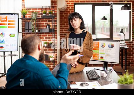 Donna manager seduto al tavolo e tenendo in mano una tazza di caffè mentre la dipendente spiega la strategia di marketing per l'azienda startup. Donna d'affari che ascolta il giovane uomo che descrive l'analisi finanziaria in un ufficio a muro. Foto Stock