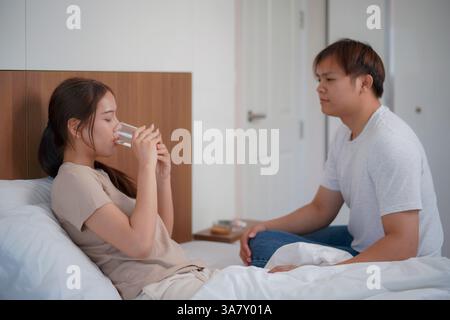 una giovane donna si siede su un letto e sta mangiando droga mentre tiene un bicchiere d'acqua. Un uomo si siede accanto a lei, offrendo rassicurazione e preoccupazione per il suo benessere. Foto Stock