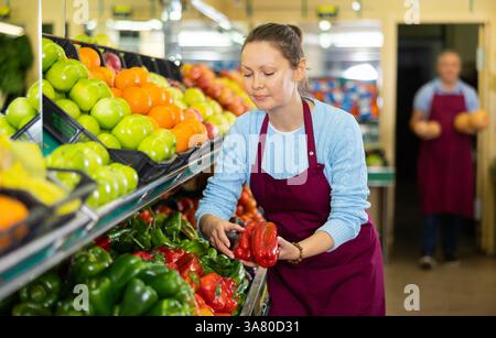 Venditore femminile adulto professionista in grembiule mettendo peperoni rossi e verdi sulle bancarelle in ipermercato Foto Stock