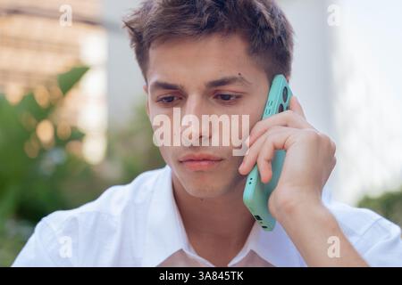 Giovane serio che parla su smartphone all'aperto con una camicia bianca alla luce del giorno Foto Stock