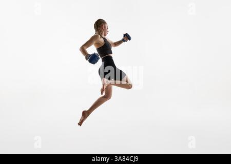 Donna con trecce catturate in salto alto con guanti da boxe, feroce allenamento focale su sfondo bianco. Foto Stock