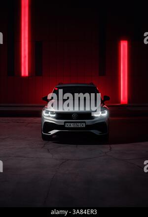 VW Tiguan argento di notte vicino a un muro con strisce rosse brillanti. Immagine verticale - Vista frontale del SUV Volkswagen su una strada buia con luci rosse. Foto Stock