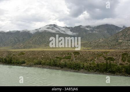 Alta sponda ripida di un ampio fiume che scorre circondato da cime di montagna sotto un cielo estivo nuvoloso. Karun, Altai, Siberia, Russia. Foto Stock