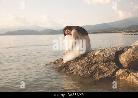 Donna in abito bianco seduta su una roccia, con uno sguardo romantico verso il mare Foto Stock